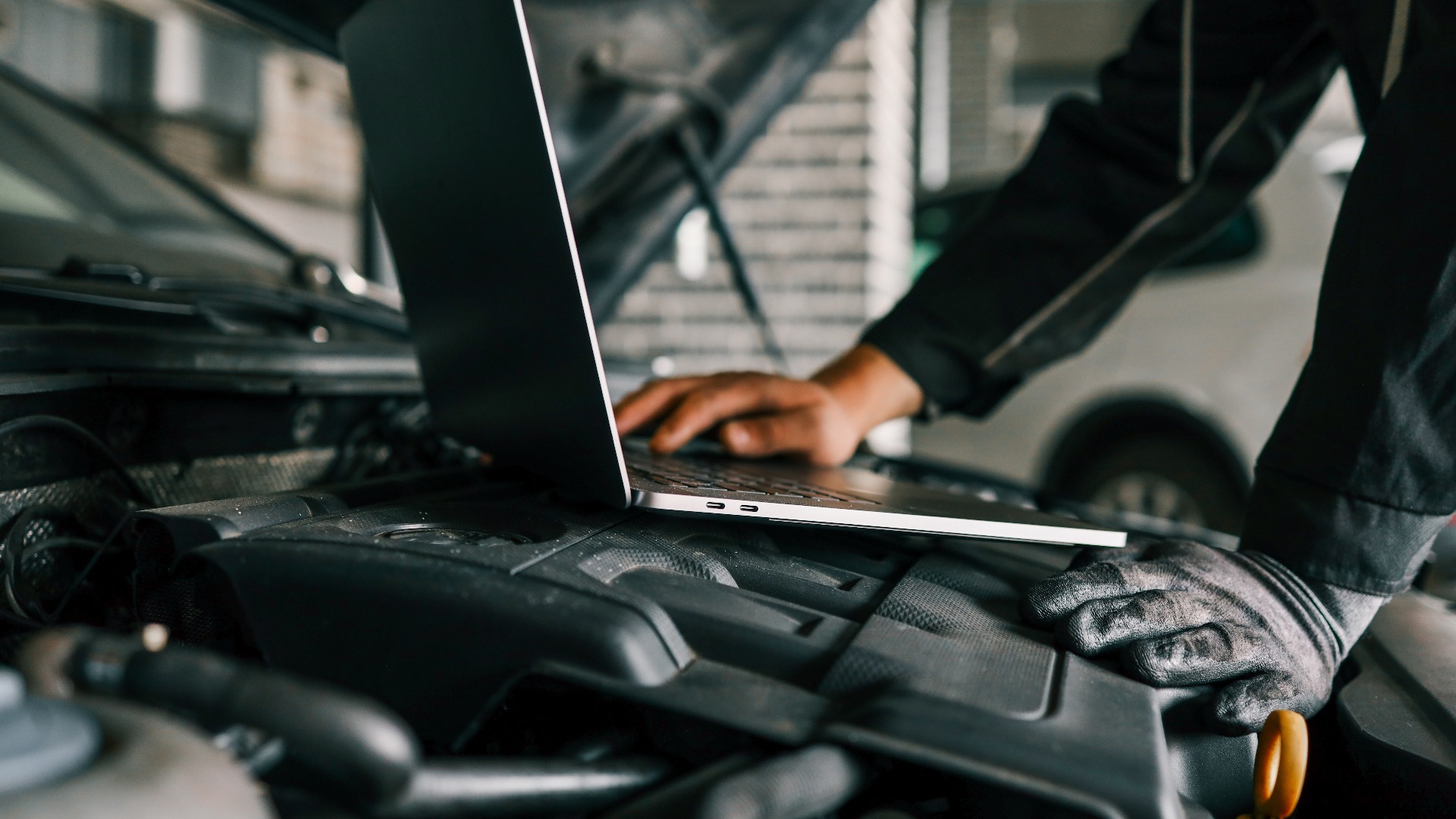 Ein Kfz-Mechaniker, der in einer Werkstatt einen Laptop zur Diagnose an einem Automotor einsetzt. Dies verdeutlicht den Einsatz von Technologie in der modernen Fahrzeugwartung und -reparatur.